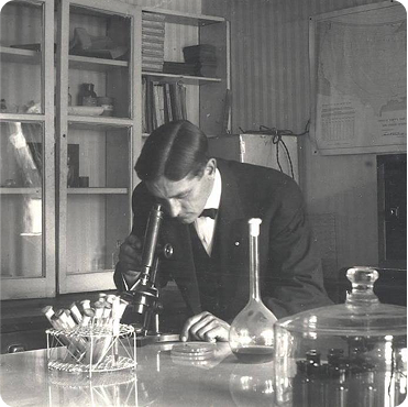 A scientist in a laboratory examines a sample through a microscope, surrounded by glassware and test tubes.