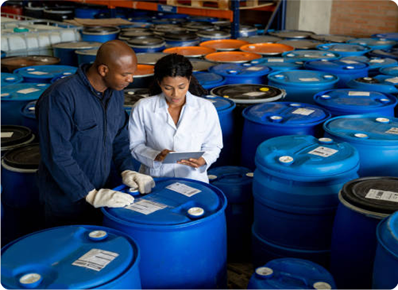 Two workers in a chemical storage area examine labels on large blue barrels, highlighting the custom manufacturing process.