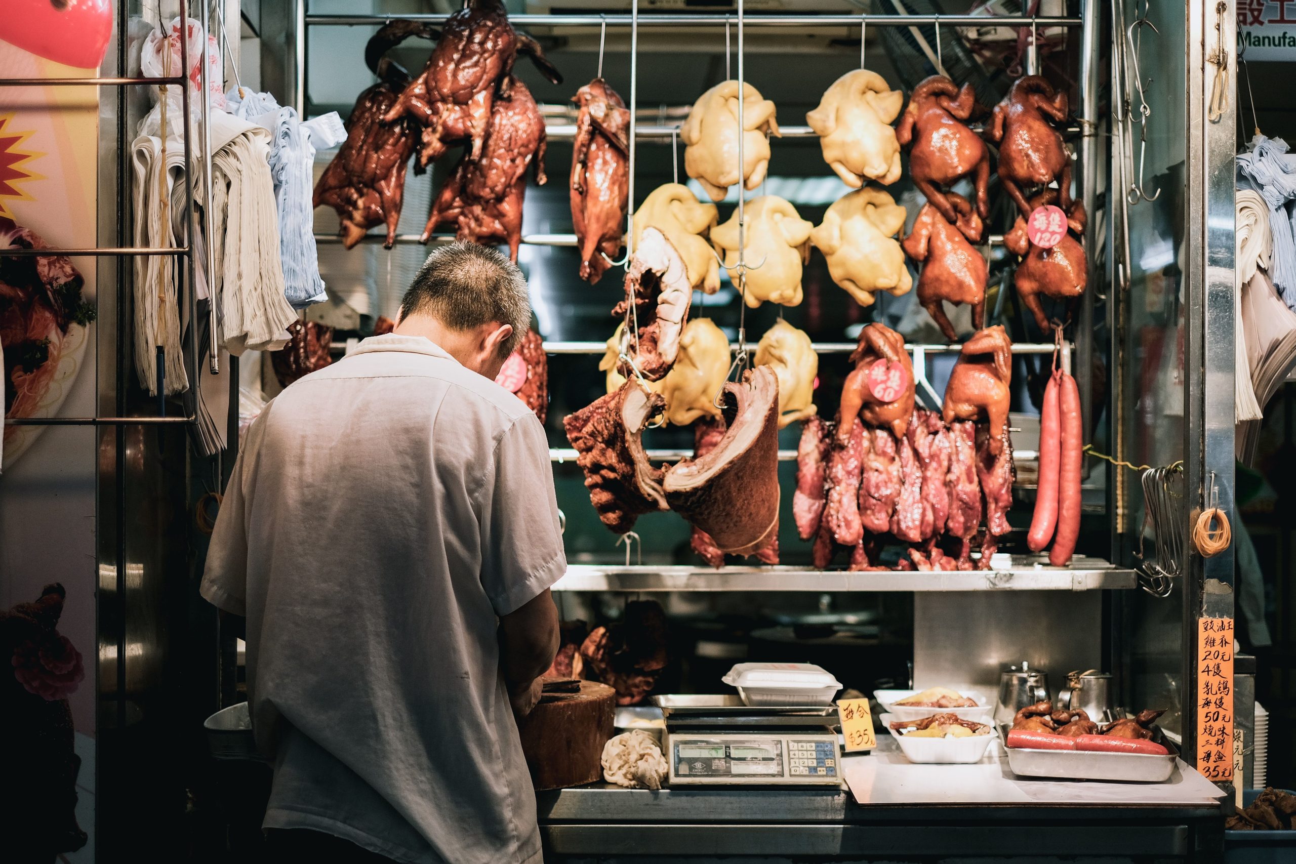 Butcher preparing meat in a shop with various cuts of poultry and sausages hanging in the background.