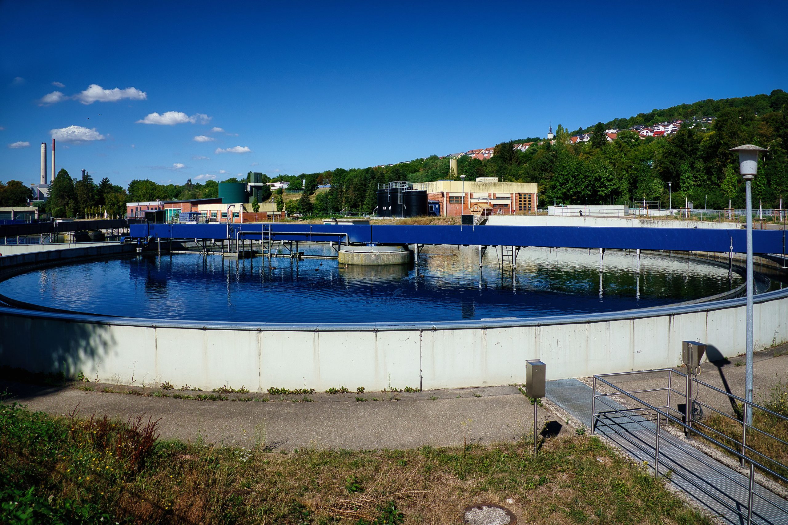Wastewater treatment facility with circular clarifier tanks surrounded by greenery and industrial buildings under a clear blue sky.