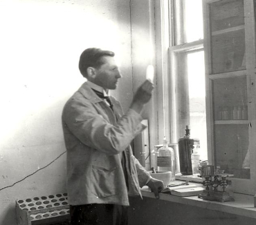 Scientist conducting bench testing in a laboratory, examining a test tube near a window with various equipment and bottles on the counter.