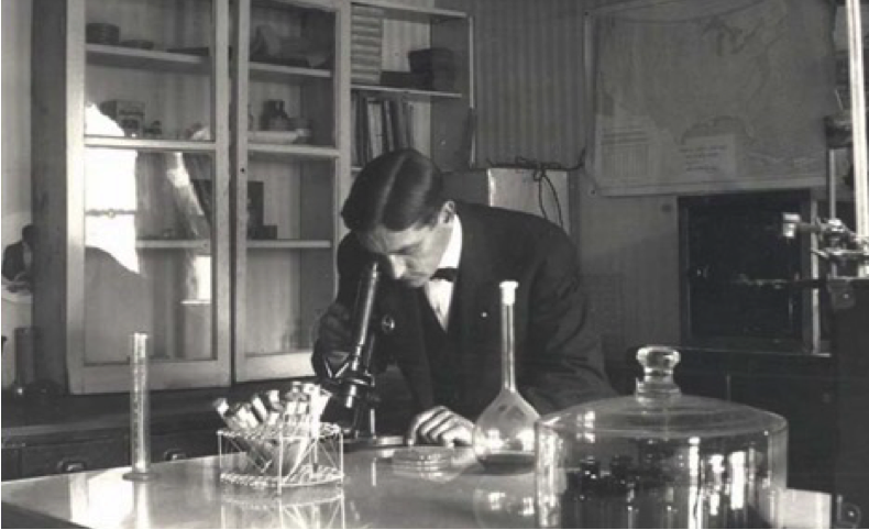 Scientist examining samples under a microscope in a laboratory setting, surrounded by glassware and scientific equipment.