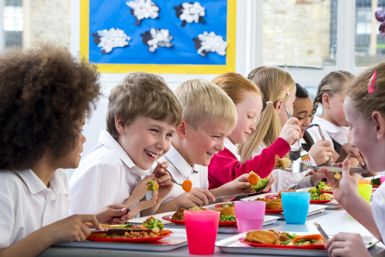 School children smiling and eating lunch together at a cafeteria table.