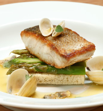 Grilled fish filet served on asparagus and bread, garnished with clams and herbs on a white plate.