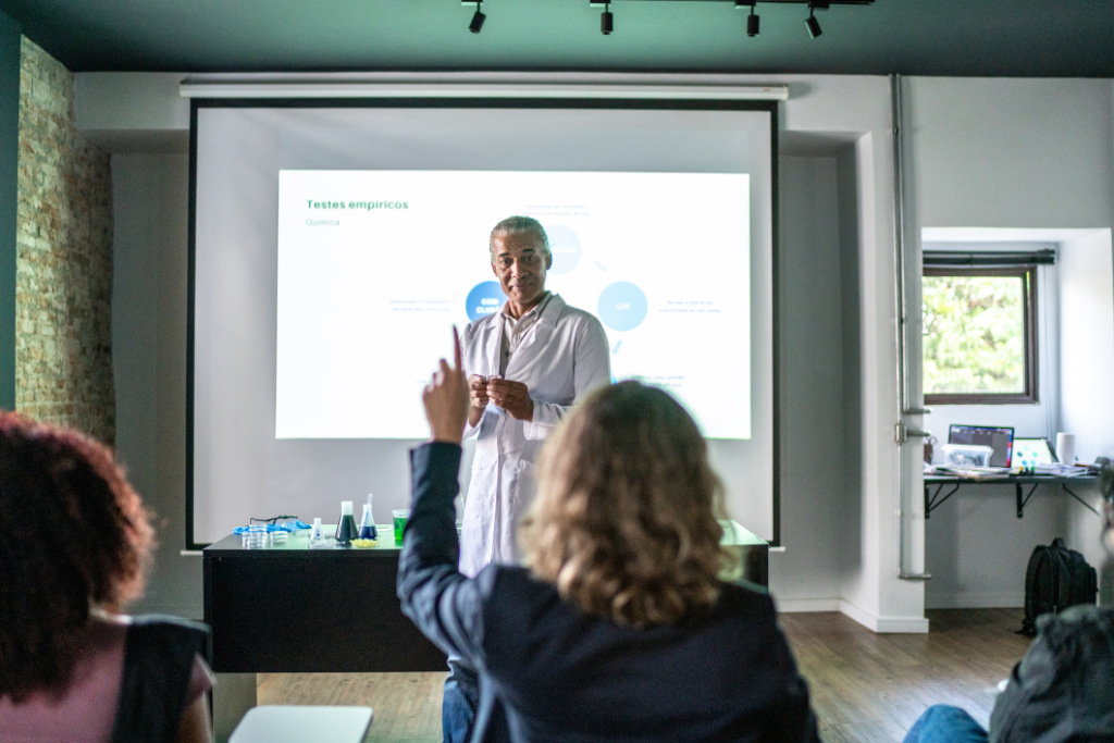 Teacher presenting in a chemistry class with a student raising their hand, in front of a projection screen displaying a diagram.