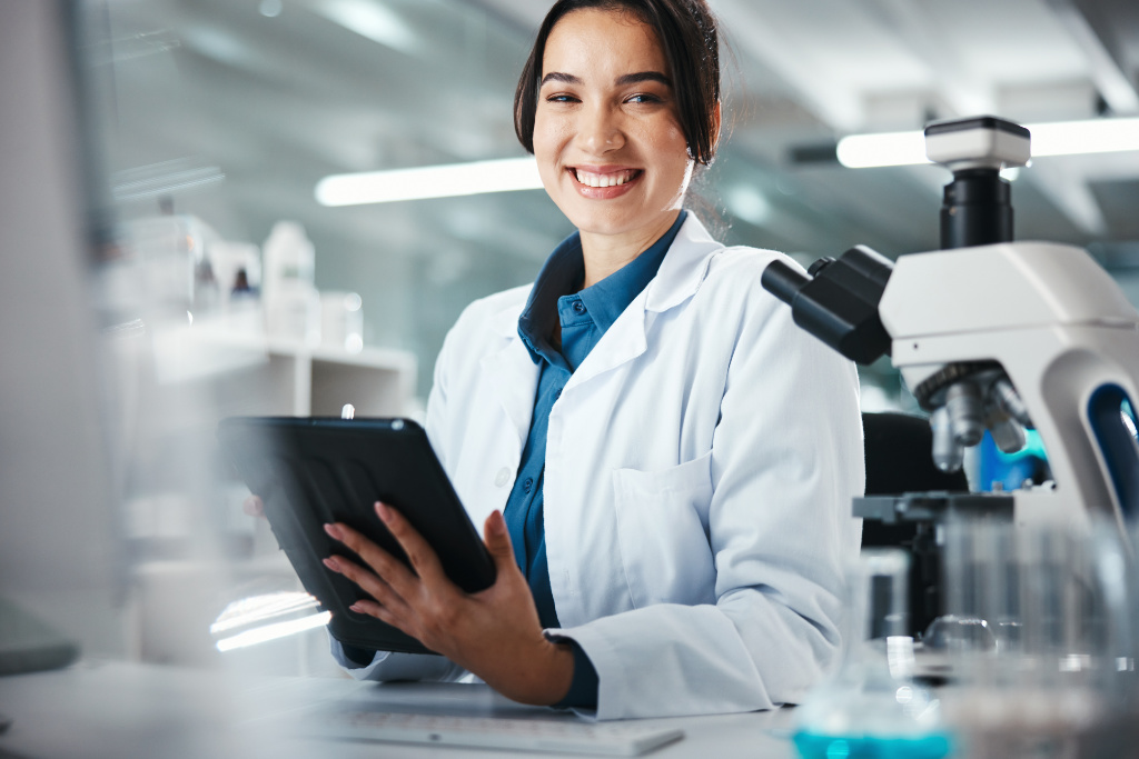 Scientist smiling while holding a tablet in a laboratory, with a microscope in the foreground, illustrating research and biotechnology.