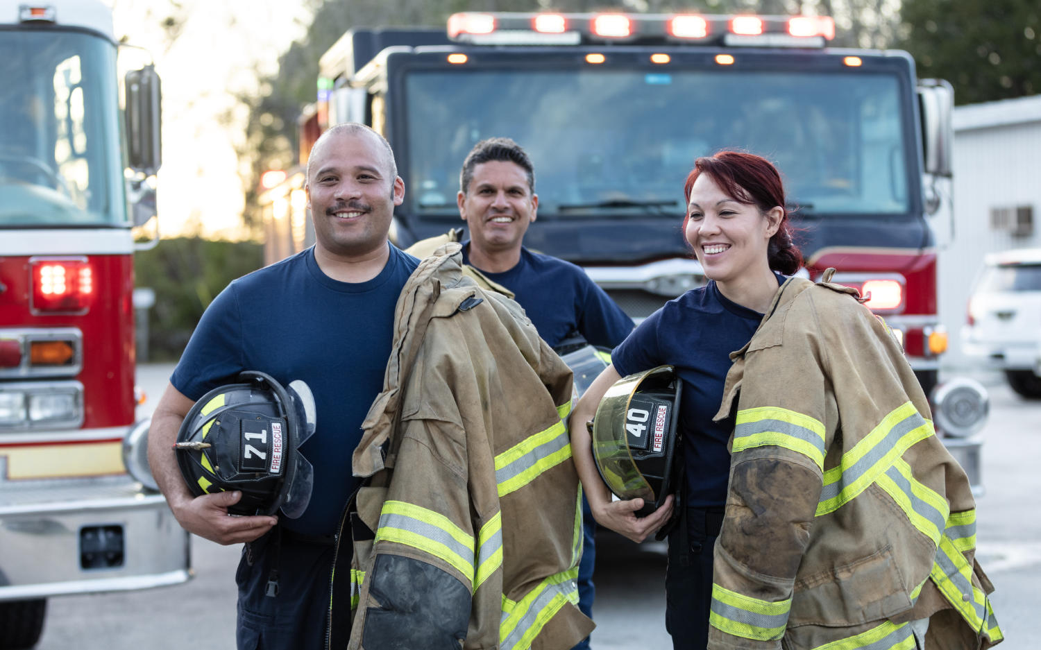 Firefighters carrying helmets and protective suits, standing in front of fire engines, smiling and appearing ready for action.