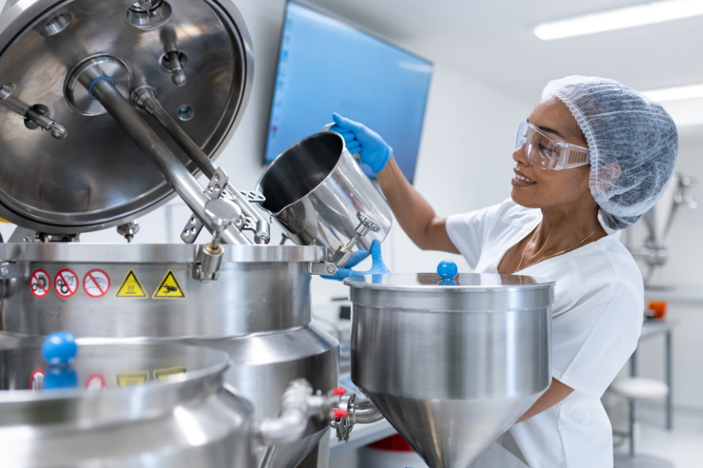 Worker in a white protective suit pours liquid into a homogenizer in a pharmaceutical manufacturing facility.