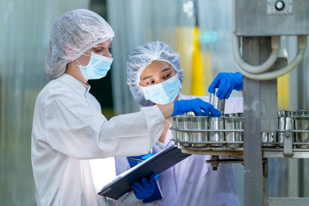 Quality control officers in a canned food manufacturing factory inspect and sample products on an automated conveyor belt.