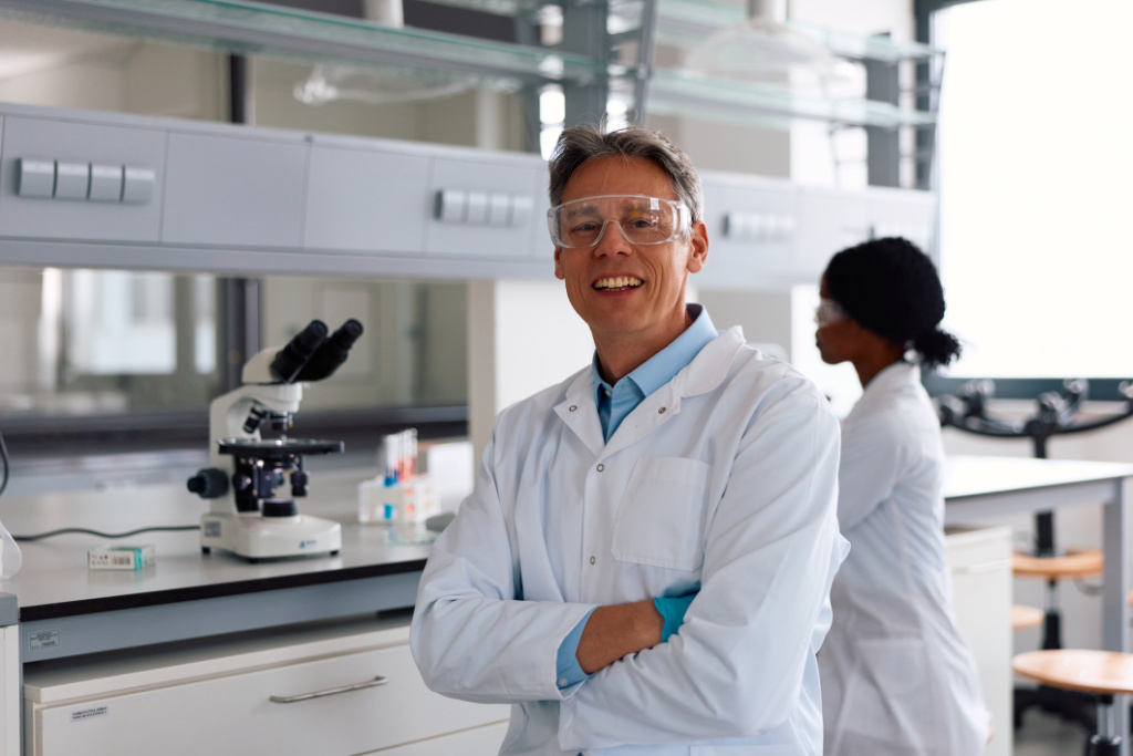 Scientist wearing safety goggles and a lab coat, smiling with arms crossed in a modern research laboratory, with a colleague working in the background.