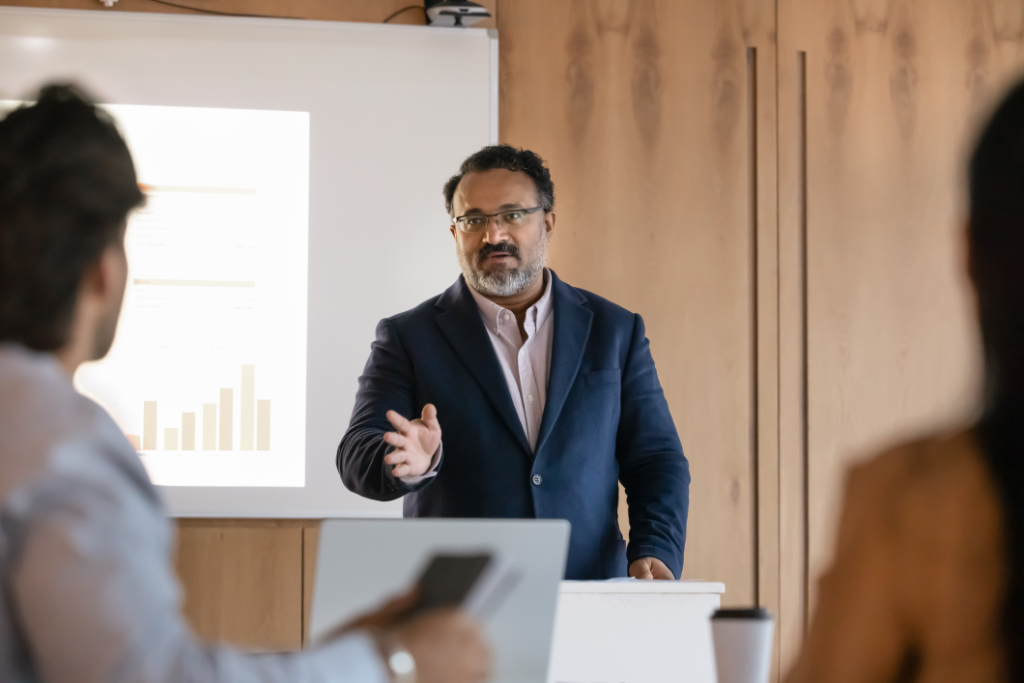 Business professional giving a presentation to a group, standing at a podium with a projected slide showing a bar graph in the background.