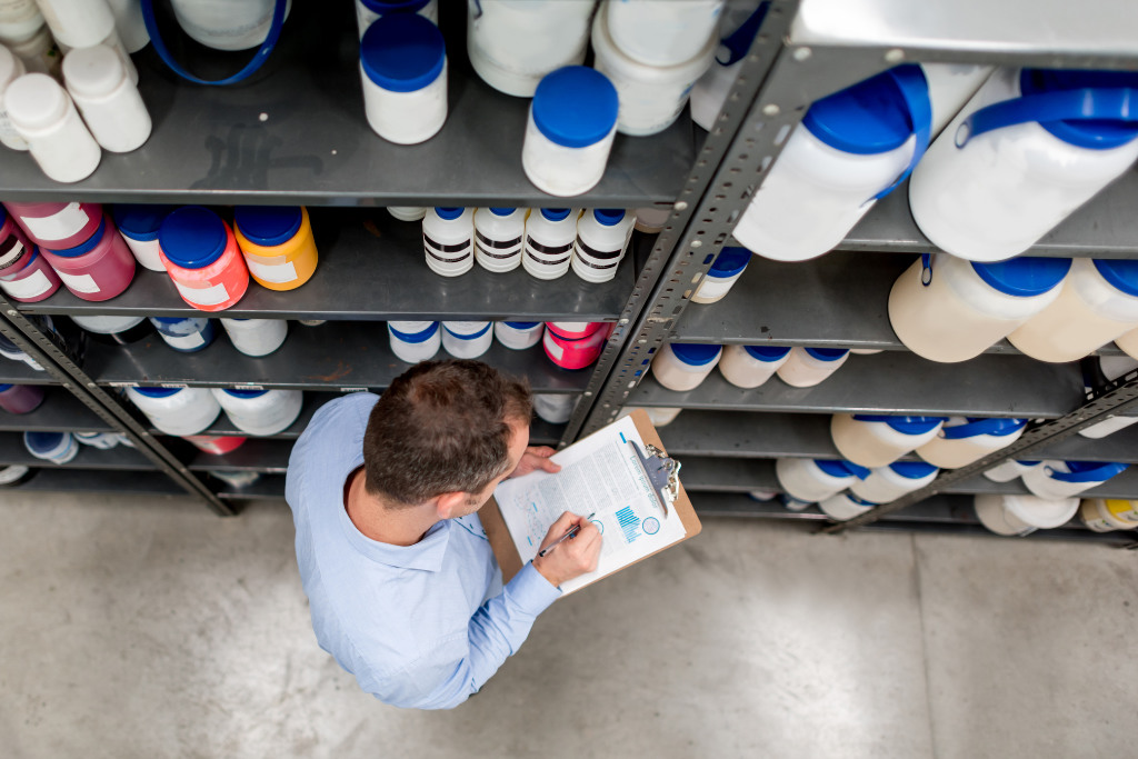 Worker conducting inventory in a chemical warehouse, writing on a clipboard surrounded by shelves of labeled containers.