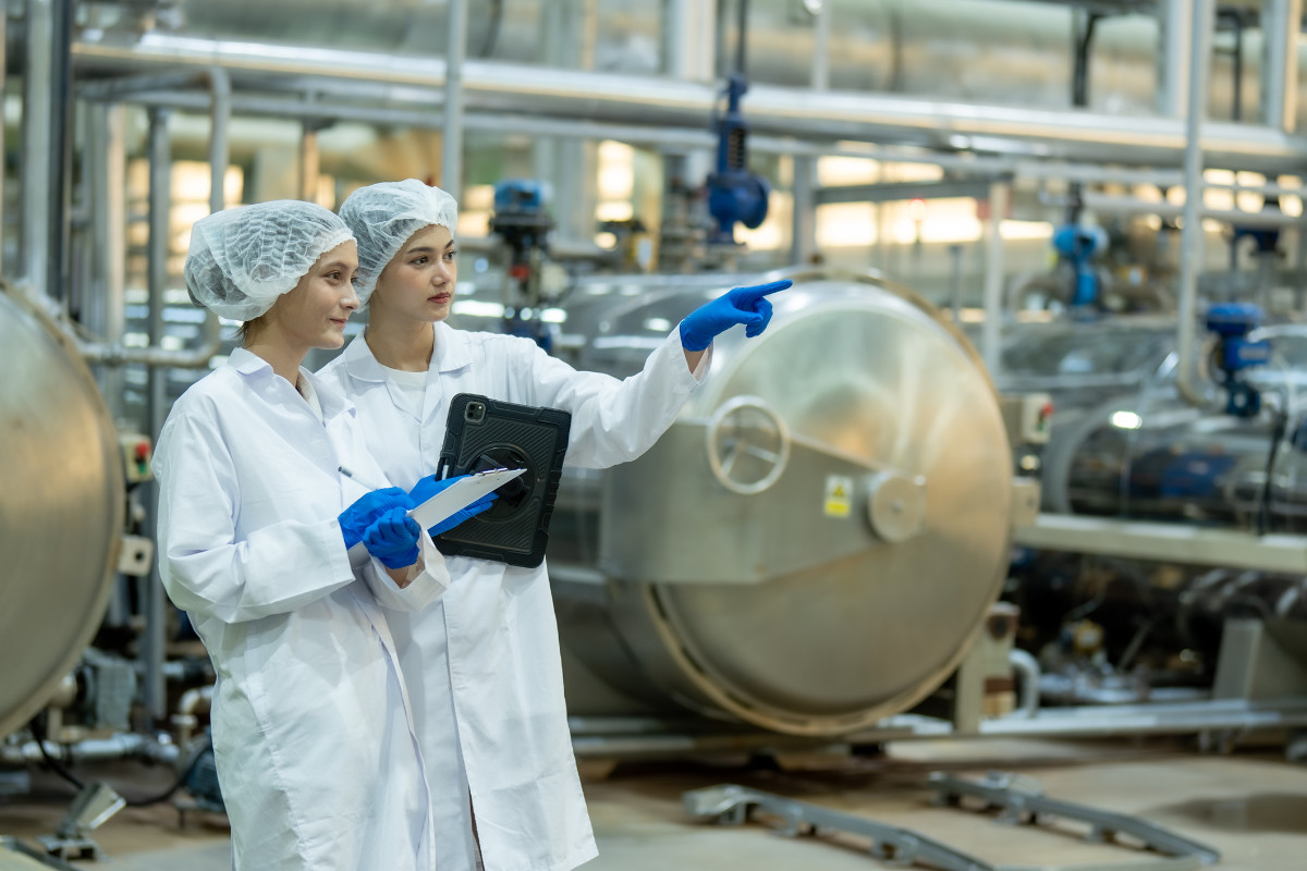 Food industry professionals wearing hair nets and sterile clothing inspect beverage production equipment, emphasizing quality control and sanitation.